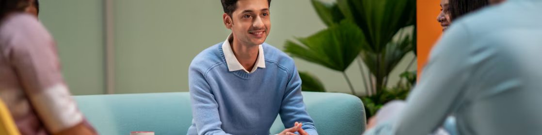 A person in a light blue sweater is seated in a lounge area, listening attentively to others. The setting includes colorful chairs, coffee mugs, and a relaxed atmosphere suitable for brainstorming or informal meetings.