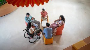 An overhead view of a small group discussion in a modern lounge area. The group is seated around a white table and chairs, including one person in a wheelchair. The space is open and well-lit, with a contemporary design and a large decorative element hanging above.