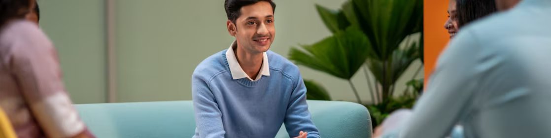 A person in a light blue sweater is seated in a lounge area, listening attentively to others. The setting includes colorful chairs, coffee mugs, and a relaxed atmosphere suitable for brainstorming or informal meetings.