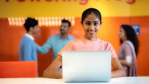 An individual is seated at a desk working on a laptop. The background shows the same orange wall with “Powering Progress” text and a group of people conversing, reinforcing the collaborative and energetic environment.