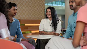A small group is gathered around a white table in a lounge area. The setting includes patterned walls and modern furniture. One person is physically specially abled, and the group seems to be engaged in an informal discussion.​