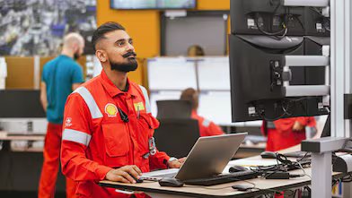 A professional in a red uniform with a badge and lanyard is focused on work at a laptop