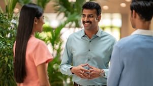 A person in a light green button-down shirt is engaged in conversation with two others. The setting appears to be an indoor space with lush green plants in the background, giving a natural and welcoming atmosphere. The individual in the center is using hand gestures, suggesting an active discussion or explanation.​
