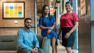 Three professionals with dark hair smiling and posing for a photo in an office. A man in a light blue shirt and glasses sits on a sofa, with two women standing beside him—one in a blue dress and the other in a red top and black skirt.