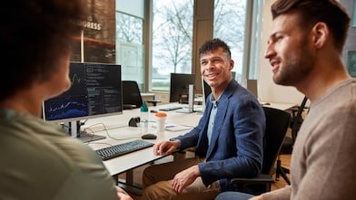 Three diverse people conversing in an office, with a computer monitor displaying graphs and codes in front of them. A man with dark curly hair in a blue blazer smiles at an out-of-focus woman with dark hair, while another man looks at her and appears to be engaged.