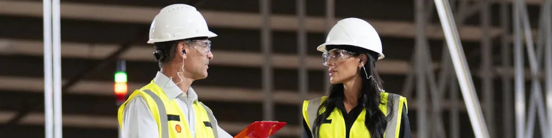 Two Shell emplyees wearing hardhats and yellow reflective jacket interacting in warehouse environment.